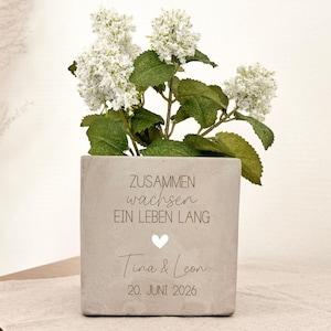 Könnte beinhalten: Ein quadratischer, hellgrauer Pflanztopf mit weißen Blüten und grünen Blättern. Der Topf trägt den Text "ZUSAMMEN wachsen EIN LEBEN LANG" und die Namen "Tina & Leon" mit einem Datum. Der Topf steht auf einer beigen Oberfläche.
