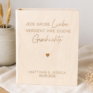 Könnte beinhalten: Ein Fotoalbum oder Buch aus hellem Holz mit der eingravierten Aufschrift JEDE GROBE LIEBE VERDIENT IHRE EIGENE GESCHICHTE, einem kleinen Herzsymbol sowie den Namen MATTHIAS & JESSICA und dem Datum 05.09.2026 auf dem vorderen Einband.