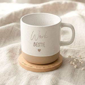 Könnte beinhalten: Ein weißer Keramikbecher mit gesprenkelter Oberfläche und beigem Boden, bedruckt mit dem Text "Work BESTIE" und einem kleinen Herzsymbol. Der Becher steht auf einem runden Holzuntersetzer mit der Gravur "DU BIST ETWAS GANZ BESONDERES".
