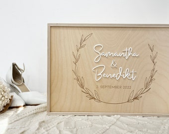 Könnte beinhalten: Eine Holzkiste mit hellbrauner Oberfläche. Die Kiste trägt eine weiße Aufschrift mit dem Text "Samantha & Benedikt 3. September 2022" umgeben von einem floralen Design.