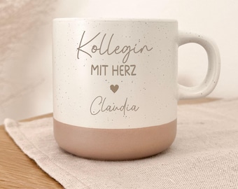 Può includere: Una tazza in ceramica con una sezione superiore color crema maculata e una base marrone chiaro a tinta unita. Il testo Kollegin MIT HERZ e il nome Claudia sono stampati in grigio scuro, separati da una piccola icona a forma di cuore.