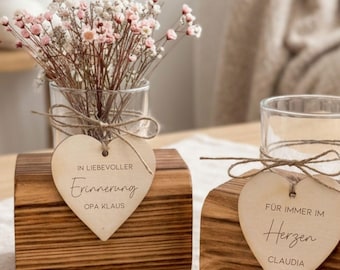 Puede incluir: Dos jarrones de bloque de madera con inserciones de vidrio, cada uno con una etiqueta de madera en forma de corazón atada con cordel. Una etiqueta dice IN LIEVOLLER Erinnerung OPA KLAUS, la otra FÜR IMMER IM Herzen CLAUDIA. Flores secas.