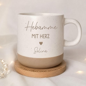 Könnte beinhalten: Ein gesprenkelter weißer Keramikbecher mit beigem Boden und Henkel. Der Text lautet "Hebamme MIT HERZ" mit einem kleinen Herzsymbol und dem Namen "Selina" darunter. Der Becher steht auf einem runden Holzuntersetzer mit der Gravur "DU BIST ETWAS GANZ BESONDERES."