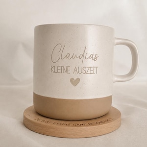Könnte beinhalten: Ein zweifarbiger Keramikbecher mit gesprenkeltem, cremeweißem Oberteil und beigefarbenem Boden. Der Becher trägt die Aufschrift "Claudias KLEINE AUSZEIT" und ein kleines Herzsymbol. Er steht auf einem runden Holzuntersetzer mit der Gravur "DU BIST ETWAS GANZ BESONDERES."