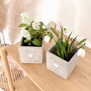 May include: Two square, light gray concrete planters with white flowers and green leaves. One planter says "Oma, Du bist wunderbar" and the other says "Mama, Du bist wunderbar" in script. Both have a white heart.