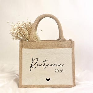 Könnte beinhalten: Eine rechteckige Jute-Tragetasche mit zwei Henkeln und einer cremefarbenen Frontpartie aus Canvas. Auf der Vorderseite ist der schwarze Schriftzug "Rentnerin 2026" über einem kleinen schwarzen Herzsymbol aufgedruckt. Weiße Trockenblumen stecken in der Tasche.