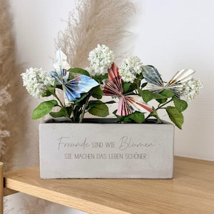Könnte beinhalten: Ein rechteckiger Beton-Pflanzkübel mit dem Text "Freunde sind wie Blumen, Sie machen das Leben schöner." enthält weiße Blüten und Papierfalter in Blau-, Rot- und Beigetönen. Der Pflanzkübel steht auf einer Holzoberfläche.