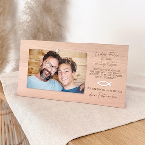 Könnte beinhalten: Ein hellfarbiger Holzbilderrahmen mit einem Foto von zwei Personen. Der Rahmen enthält deutschen Text, darunter "Lieber Felix" und "Zur Konfirmation, am 18. Mai 2026". Eine kleine silberne Nadel sichert das Foto. Der Rahmen liegt auf einem beigen Tuch.