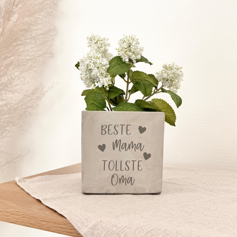 K&ouml;nnte beinhalten: Ein quadratischer, hellgrauer Beton-Blumentopf mit den Worten "BESTE Mama TOLLSTE Oma" und Herzsymbolen. Der Topf enth&auml;lt wei&szlig;e Blumen mit gr&uuml;nen Bl&auml;ttern. Der Topf steht auf einem beigen Leinentuch.