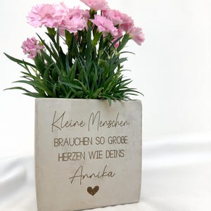 Op de afbeelding: Een vierkante, lichtgrijze plantenbak met roze anjers en groene bladeren. De plantenbak heeft de gegraveerde tekst "Kleine Menschen brauchen so grobe Herzen wie Deins Annika" met een hartsymbool.
