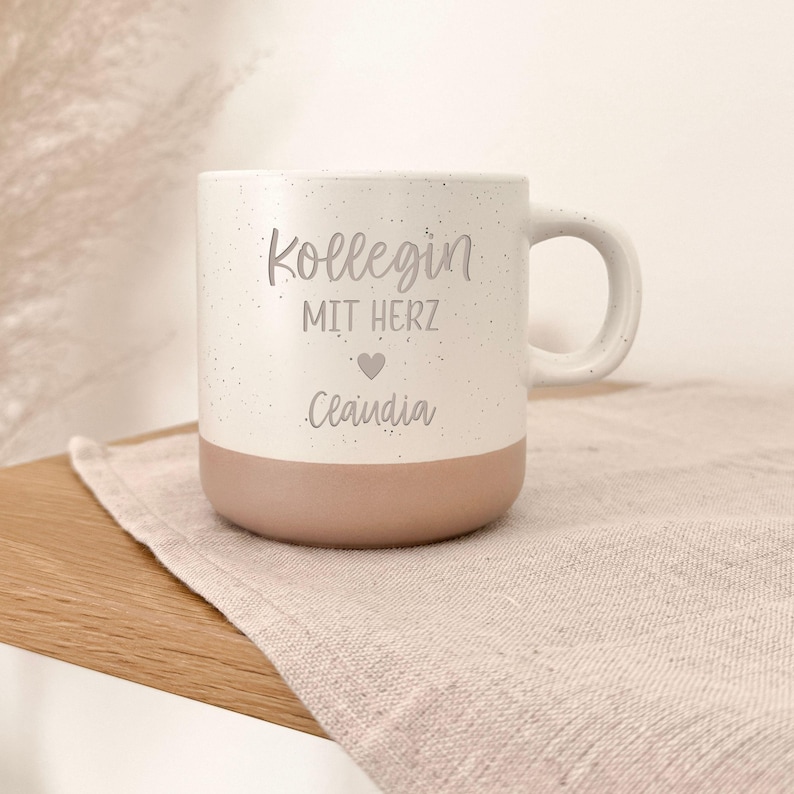 May include: White ceramic mug with a speckled finish and a light brown base. The mug features the text "Kollegin MIT HERZ Claudia" in a gray font, with a heart symbol. The mug has a curved handle and is sitting on a beige cloth.