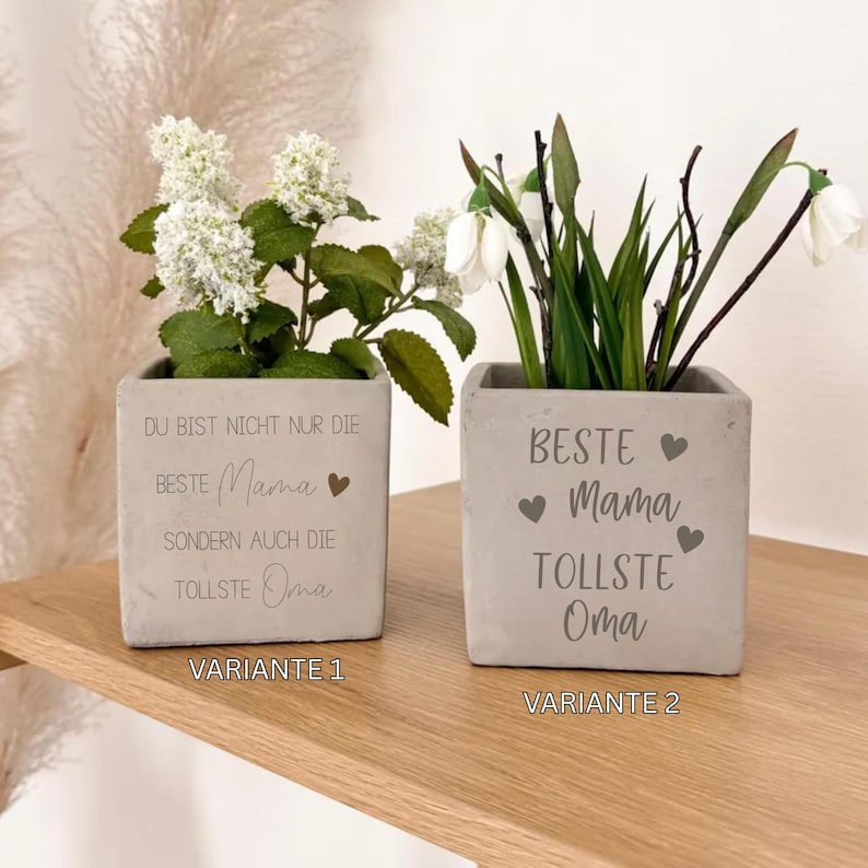 K&ouml;nnte beinhalten: Zwei quadratische, graue Betonpflanzgef&auml;&szlig;e mit wei&szlig;en Blumen. Ein Pflanzgef&auml;&szlig; hat den Text "DU BIST NICHT NUR DIE BESTE Mama SONDERN AUCH DIE TOLLSTE Oma" und das andere "BESTE Mama TOLLSTE Oma".