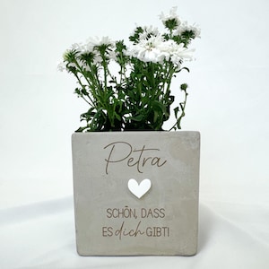 Könnte beinhalten: Ein quadratischer, hellgrauer Beton-Pflanztopf mit weißen Blumen. Der Topf trägt den Namen "Petra" in Schreibschrift, ein weißes Herz und den deutschen Text "SCHÖN, DASS ES dich GIBT!" Die Blumen haben weiße Blütenblätter und grüne Stiele.