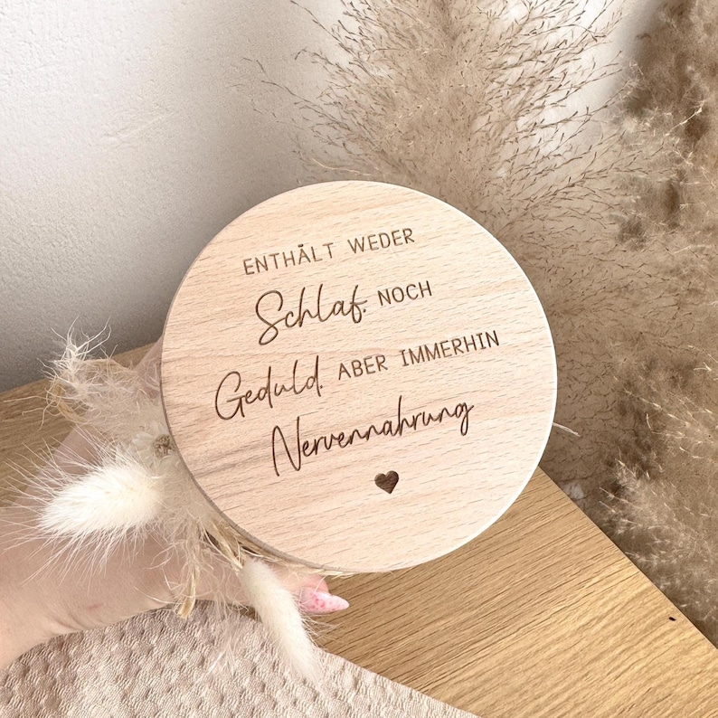 May include: A round wooden sign with engraved text in German. The text reads "ENTH&Auml;LT WEDER Schlaf NOCH Geduld ABER IMMERHIN Nervennahrung" with a small heart symbol. The sign is light brown and held in a hand, with dried flowers in the background.