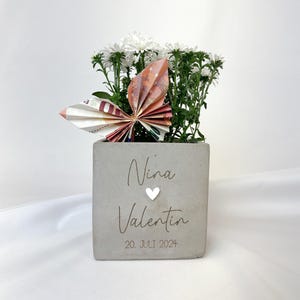 Könnte beinhalten: Ein grauer Beton-Pflanztopf mit weißen Blumen und einem Schmetterling aus gefalteten Geldscheinen. Der Topf ist mit den Namen "Nina" und "Valentin", einem Herz und dem Datum "20. JULI 2024" graviert.