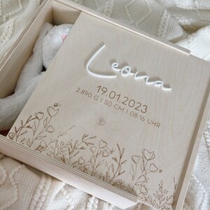 Può includere: Una scatola di legno per ricordi con un design floreale inciso sul coperchio. Il coperchio è aperto e all'interno c'è un peluche bianco. La scatola è personalizzata con il nome "Leona" e la data "19.01.2023" insieme all'ora, al peso e all'altezza del bambino.
