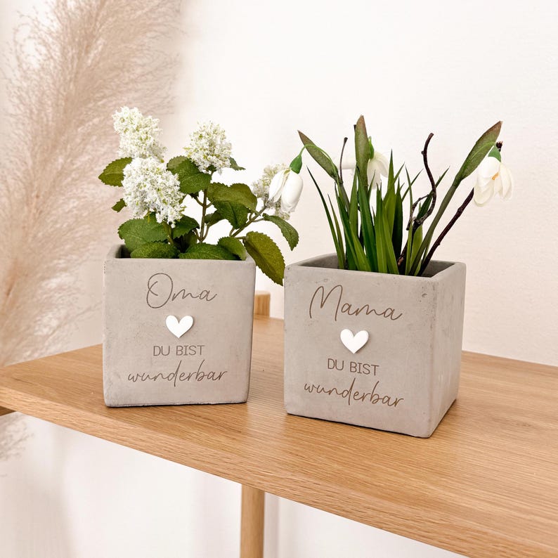 May include: Two square, gray concrete planters with white flowers and greenery. The planter on the left has the word "Oma" and the text "DU BIST wunderbar" with a white heart. The planter on the right has the word "Mama" and the same text and heart.