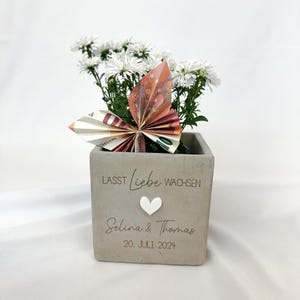 Könnte beinhalten: Ein quadratischer, grauer Beton-Blumentopf mit weißen Blumen und einem Papier-Schmetterling aus Geldscheinen. Der Topf trägt die Worte "Lasst Liebe wachsen", ein weißes Herz und die Namen "Selina & Thomas" mit dem Datum "20. JULI 2024" eingraviert.