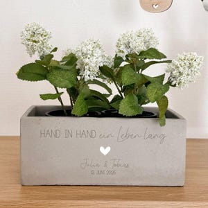 Op de afbeelding: Een rechthoekige, grijze betonnen plantenbak met witte bloemen en groene bladeren. De plantenbak heeft de tekst "HAND IN HAND ein Leben lang", een hartsymbool en de namen "Julia & Tobias" met de datum "12. JUNI 2025".