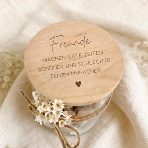 Könnte beinhalten: Ein klares Glas mit einem Holzdeckel, gebunden mit Bindfaden und verziert mit kleinen weißen Blumen. Der Deckel ist mit dem Wort "Freunde" und einem Zitat auf Deutsch graviert. Ein kleines Herz ist ebenfalls auf dem Deckel eingraviert.