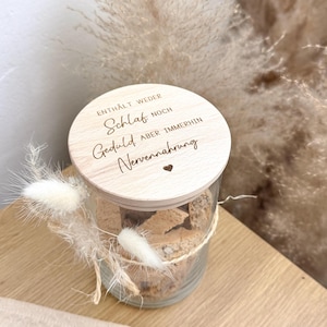 May include: A clear glass jar with a wooden lid, containing a light brown substance. The lid is engraved with German text and a heart symbol. The jar is decorated with dried flowers and tied with twine. The jar sits on a wooden surface.