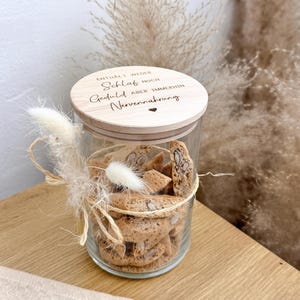May include: A clear glass jar filled with biscotti cookies, topped with a wooden lid. The lid has engraved text and a heart symbol. The jar is decorated with dried flowers and twine, creating a rustic aesthetic.