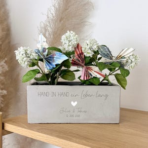 K&ouml;nnte beinhalten: Ein rechteckiger Betonpflanztopf mit wei&szlig;en Bl&uuml;ten und Origami-Schmetterlingen aus bunten Geldscheinen. Der Pflanztopf tr&auml;gt den Text "HAND IN HAND ein leben lang" und die Namen "Julia & Tobias" mit einem Datum.