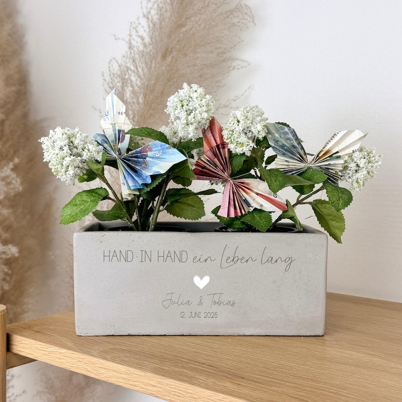 K&ouml;nnte beinhalten: Ein rechteckiger Betonpflanztopf mit wei&szlig;en Bl&uuml;ten und Origami-Schmetterlingen aus bunten Geldscheinen. Der Pflanztopf tr&auml;gt den Text "HAND IN HAND ein leben lang" und die Namen "Julia & Tobias" mit einem Datum.