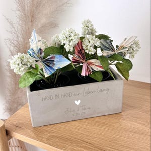 K&ouml;nnte beinhalten: Ein grauer Beton-Pflanzkasten mit wei&szlig;en Bl&uuml;ten und Origami-Schmetterlingen aus Geldscheinen. Auf dem Kasten steht der Text "HAND IN HAND ein Leben lang", ein Herz und Namen mit Datum. Der Kasten steht auf einer Holzoberfl&auml;che.
