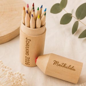 Könnte beinhalten: Ein zylindrisches Stifteetui aus hellem Holz mit verschiedenen Buntstiften. Das Etui ist mit dem Text "SCHULKIND 2026" graviert und hat einen passenden abnehmbaren Deckel, der mit dem Namen "Mathilda" in Schreibschrift graviert ist.