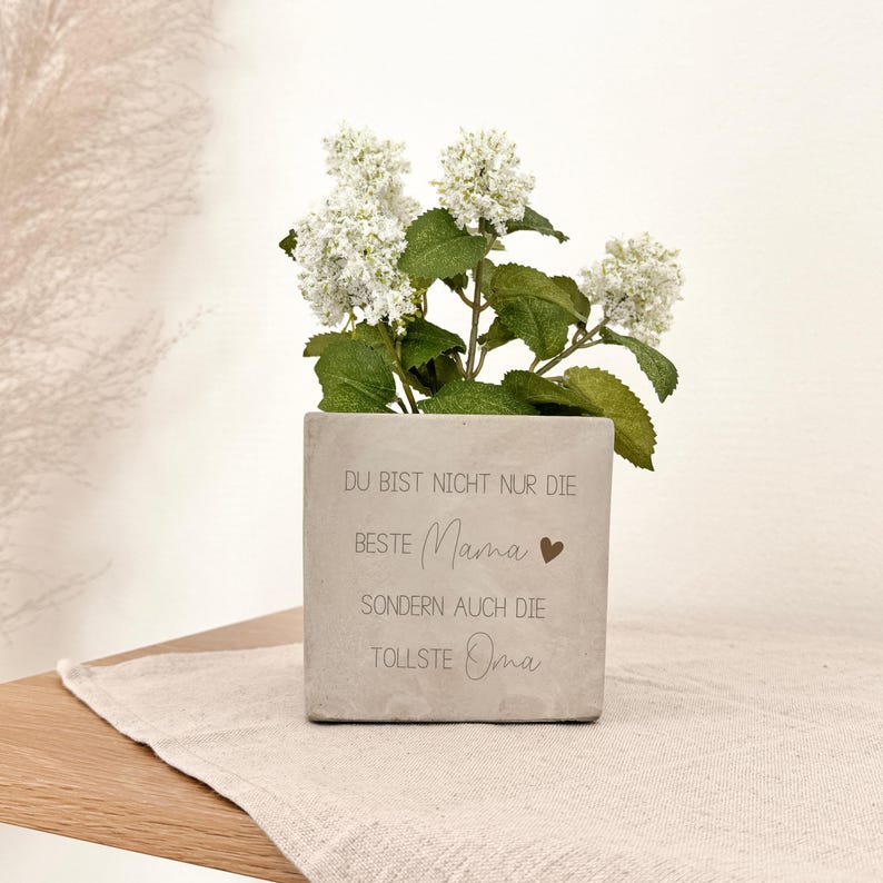 K&ouml;nnte beinhalten: Ein quadratischer, grauer Betonblumentopf mit dem Text "DU BIST NICHT NUR DIE BESTE Mama" und "SONDERN AUCH DIE TOLLSTE Oma" in einer Schriftart. Der Topf enth&auml;lt wei&szlig;e Blumen mit gr&uuml;nen Bl&auml;ttern.