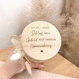 May include: A round, light-colored wooden box with the German text "ENTH&Auml;LT WEDER Schlaf NOCH Geduld ABER IMMERHIN Nervennahrung" and a small heart. The box is decorated with dried flowers and twine.