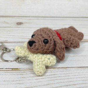 May include: Crocheted brown dog keychain with a red collar and a cream-colored bone. The dog has black eyes and a black nose. The keychain has a silver clasp and ring.