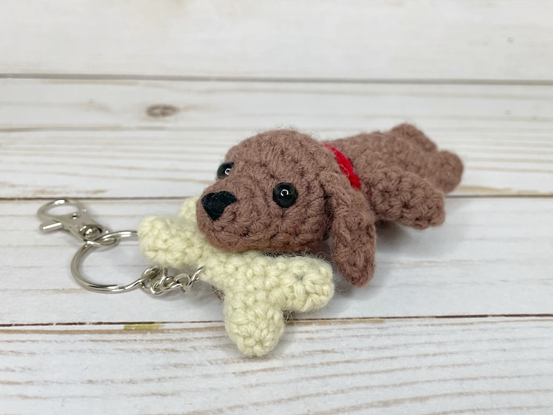 May include: Crocheted brown dog keychain with a red collar and a cream-colored bone. The dog has black eyes and a black nose. The keychain has a silver clasp and ring.