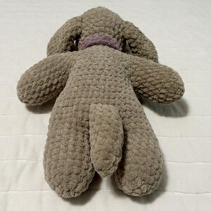 May include: A plush brown dog toy with a purple collar. The toy is made of a soft, textured fabric and has a long tail.