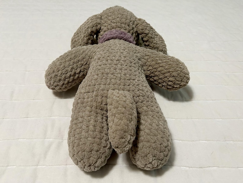 May include: A plush brown dog toy with a purple collar. The toy is made of a soft, textured fabric and has a long tail.