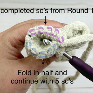 May include: A close-up of a crocheted project with a purple crochet hook. The project is folded in half and the crocheter is working on the next round of stitches. The text "5 completed sc's from Round 17" and "Fold in half and continue with 5 sc's" is visible in the image.
