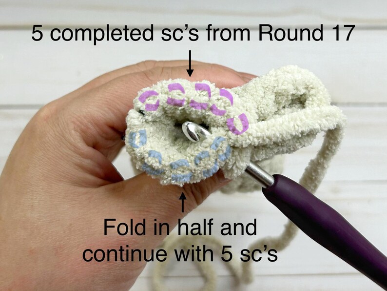 May include: A close-up of a crocheted project with a purple crochet hook. The project is folded in half and the crocheter is working on the next round of stitches. The text "5 completed sc's from Round 17" and "Fold in half and continue with 5 sc's" is visible in the image.