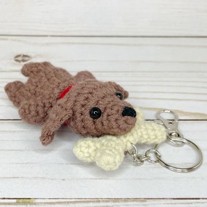 May include: A handmade crochet dog keychain in brown with a red collar and a cream-colored bone. The dog has black button eyes and a black nose. The keychain has a silver clasp and ring.