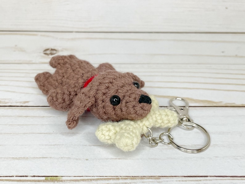 May include: A handmade crochet dog keychain in brown with a red collar and a cream-colored bone. The dog has black button eyes and a black nose. The keychain has a silver clasp and ring.