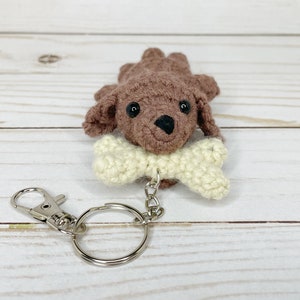 May include: A crocheted brown dog keychain with a cream-colored bone in its mouth. The dog has black eyes and a black nose. The keychain includes a silver-colored clasp and a key ring.