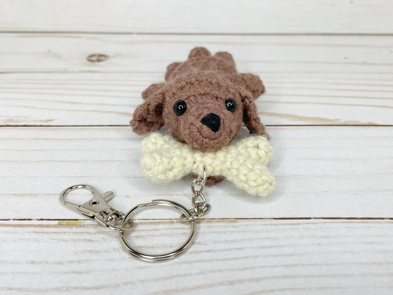 May include: A crocheted brown dog keychain with a cream-colored bone in its mouth. The dog has black eyes and a black nose. The keychain includes a silver-colored clasp and a key ring.