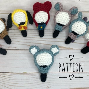 Op de afbeelding: Een verzameling van acht gehaakte amigurumi-speelgoed met verschillende dierenontwerpen. Elk speelgoed heeft een wit gezicht en een zwarte stok die aan de onderkant is bevestigd. De speelgoedjes zijn in een cirkel op een wit houten oppervlak gerangschikt. De tekst "PATTERN" is in zwart op het oppervlak geschreven.