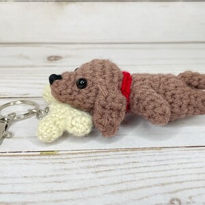 May include: A crocheted dog keychain in brown with a red collar, lying next to a cream-colored bone. The dog has black eyes and a black nose. The keychain has a silver clasp and ring.