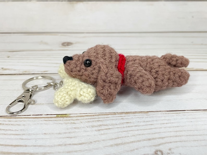 May include: A crocheted dog keychain in brown with a red collar, lying next to a cream-colored bone. The dog has black eyes and a black nose. The keychain has a silver clasp and ring.