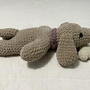 May include: A plush brown dog toy with a purple collar and a white plush tail. The toy is lying on a white surface.