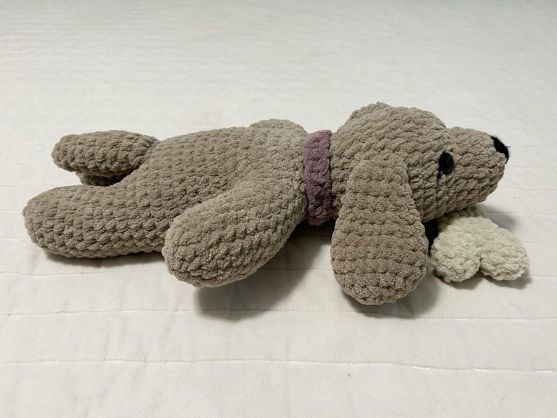 May include: A plush brown dog toy with a purple collar and a white plush tail. The toy is lying on a white surface.