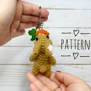 May include: A crocheted bunny keychain with a carrot on its head. The bunny is brown with pink accents and black eyes. The keychain is attached to a silver key ring.