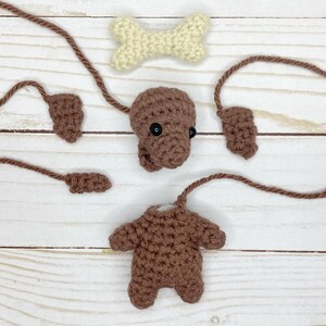 May include: A disassembled crochet dog toy with brown body parts, including a head with black eyes, ears, legs, and a tail. A cream-colored bone is also included. The pieces are arranged on a light wood surface.