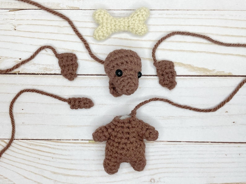 May include: A disassembled crochet dog toy with brown body parts, including a head with black eyes, ears, legs, and a tail. A cream-colored bone is also included. The pieces are arranged on a light wood surface.